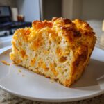Close-up of a thick slice of fluffy cheese bread, studded with melted cheddar, sitting on a white plate.
