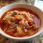 Close-up of a white bowl filled with rich, red cabbage soup featuring ground meat, rice, and chunks of cabbage.
