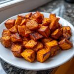 A close-up of perfectly caramelized and seasoned roasted sweet potatoes cubes piled high on a white plate.
