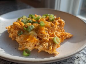 A scoop of creamy, orange buffalo chicken dip topped with sliced green onions on a light-colored plate.