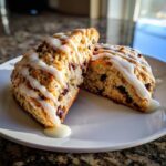 Two halves of a freshly baked chocolate chip scone drizzled with thick vanilla glaze, sitting on a white plate.