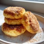 A stack of four soft, round churro cookies heavily coated in cinnamon sugar, resting on a white plate.