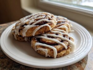 A stack of three delicious cinnamon roll cookies drizzled with white icing, sitting on a white plate.