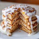 Close-up of a tall stack of cinnamon roll pancakes with a slice removed, dripping with thick white icing.