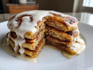 A tall stack of fluffy cinnamon roll pancakes cut open, generously drizzled with white cream cheese glaze.