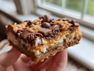 A close-up view of a thick, layered magic cookie bars slice held up, showing the crust, coconut, and melted chocolate topping.