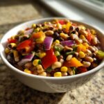 Close-up of a white bowl filled with colorful cowboy caviar recipe ingredients like black-eyed peas, corn, and bell peppers.