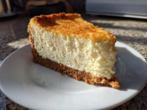 A thick, creamy slice of new york cheesecake featuring a golden-brown top and a graham cracker crust, served on a white plate.