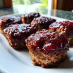 Close-up of several juicy mini meatloaf portions topped with a thick, shiny red glaze on a white plate.