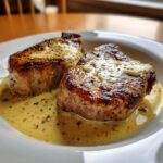 Two perfectly seared pork chops smothered in a creamy herb sauce served on a white plate.