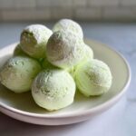 A pile of light green, round cream cheese mints dusted lightly with powdered sugar on a white plate.