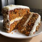 Close-up of a thick slice of moist carrot cake layered with cream cheese frosting and covered in chopped nuts.