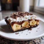 A decadent slice of no-bake banoffee pie featuring a dark crust, bananas, caramel, whipped cream, and chocolate shavings.