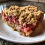 Close-up of a square serving of rhubarb crisp showing bright pink filling and thick, buttery oat crumble topping.