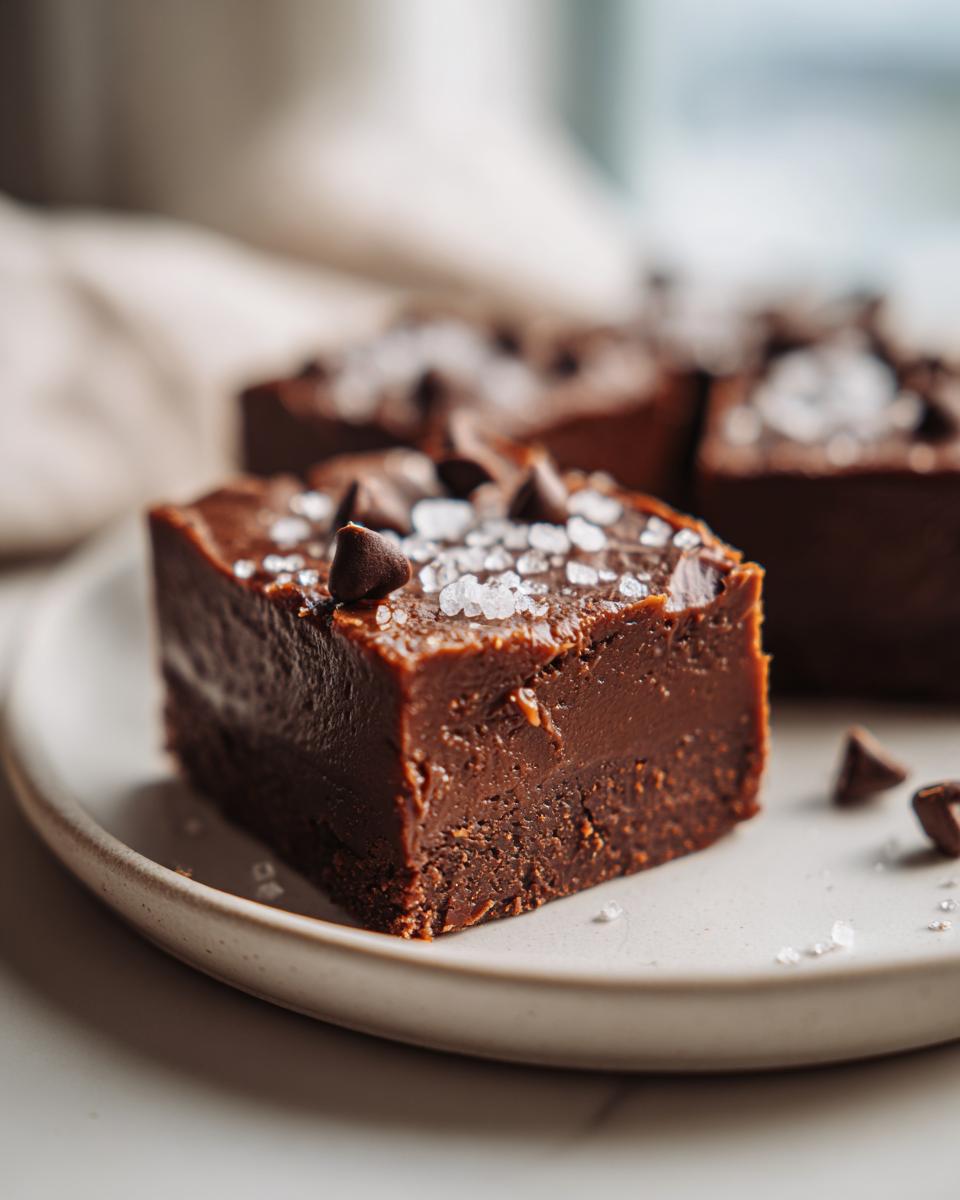 A close-up of a rich, dark chocolate salted caramel fudge square topped with sea salt flakes and chocolate chips.
