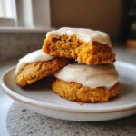 A stack of three soft, orange pumpkin cookie treats topped with thick cream cheese frosting. One cookie is broken open.