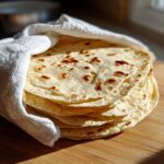A warm stack of freshly made corn tortillas wrapped partially in a white cloth on a wooden surface.