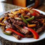 Close-up of sizzling steak fajita strips mixed with colorful sautéed bell peppers and onions on a white plate.