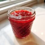 Close-up of vibrant red strawberry freezer jam filling a quilted glass jar, sitting on a white surface near a window.
