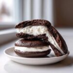 Two decadent chocolate whoopie pies stacked on a white plate, one cut in half showing the thick white cream filling.