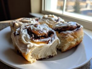 Four fluffy, freshly baked chocolate cinnamon rolls generously topped with white icing, resting on a white plate near a window.