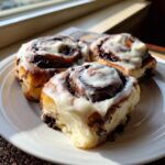 Three freshly baked chocolate cinnamon rolls generously topped with white icing, sitting on a white plate near a window.