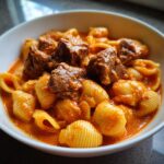 A close-up of a white bowl filled with creamy paprika steak and shells pasta in a rich, orange sauce.