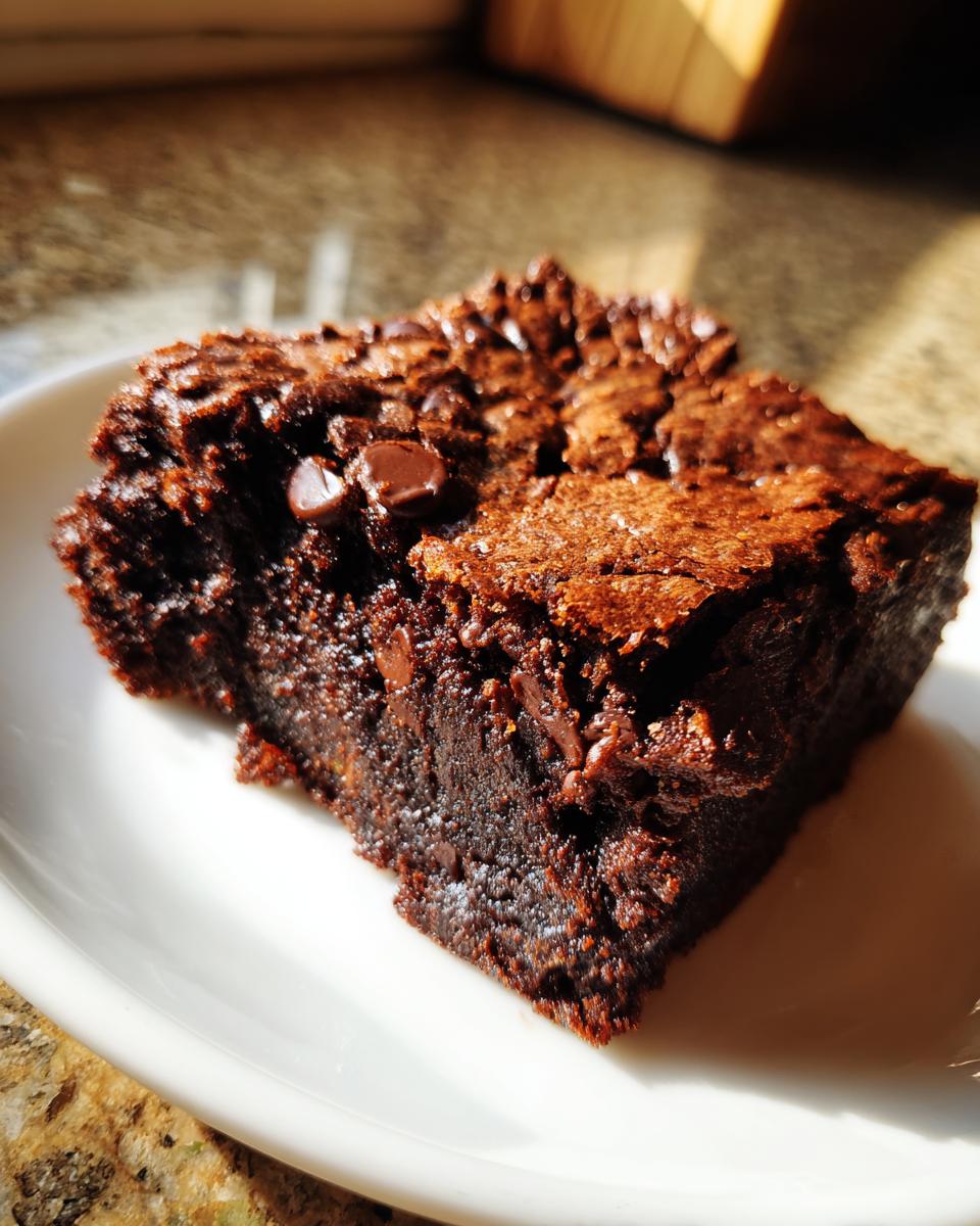 Close-up of a rich, fudgy square of zucchini brownies topped with melted chocolate chips, sitting on a white plate.