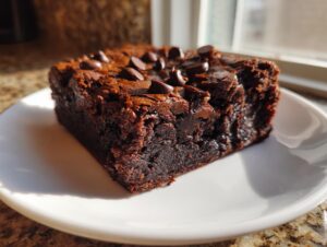 A close-up of a rich, fudgy zucchini brownies slice topped with melted chocolate chips on a white plate.