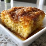 Close-up of a warm, gooey slice of snickerdoodle cobbler with a cinnamon-sugar topping in a white dish.
