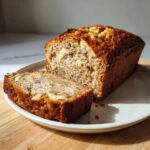 A loaf of moist greek yogurt banana bread, partially sliced, resting on a white plate.