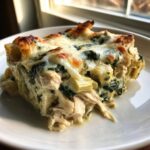 A close-up of a cheesy serving of spinach artichoke chicken casserole on a white plate.