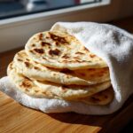 A stack of warm, freshly made greek pita bread wrapped partially in a white towel on a wooden surface.